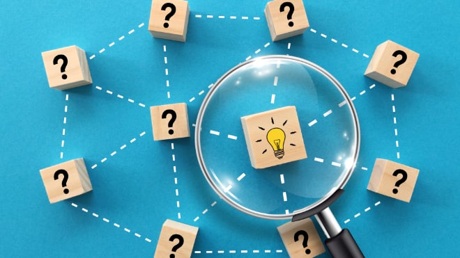A magnifying glass inspecting a number of wooden cubes with question marks upon them laid upon a blue table. The wooden cube under the magnifying glass has a lightbulb painted on it.